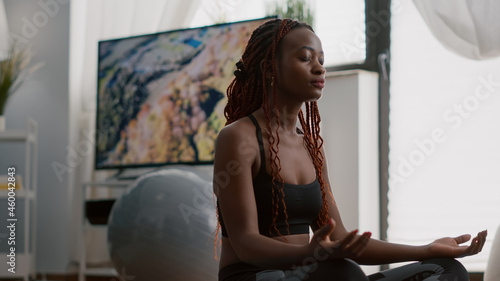 Athlete woman with black skin in sportwear practicing yoga at home in living room. Flexible african adult meditating in lotus position with closed eyes on sports mat enjoying healthy lifestyle photo