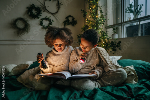 Brother and sister sitting in the bed and reading book with flashlamps photo