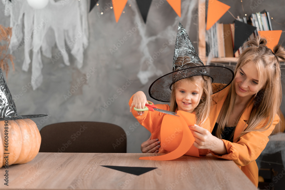 Preparing for the holiday Halloween woman and child daughter sit at home at the table wearing a witch hat and cut out paper crafts.