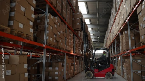 Warehouse loader standing at rack, lifting goods in boxes up and setting high on empty space. Storehouse machinery during work, forklift driver placing huge pallet of cardboard boxes on rack