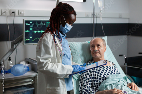 African american medical practitoner wearing chirurgical mask, in hospital room during consultation of sick unwell senior man, breathing with help from test tube and iv drip attached. photo