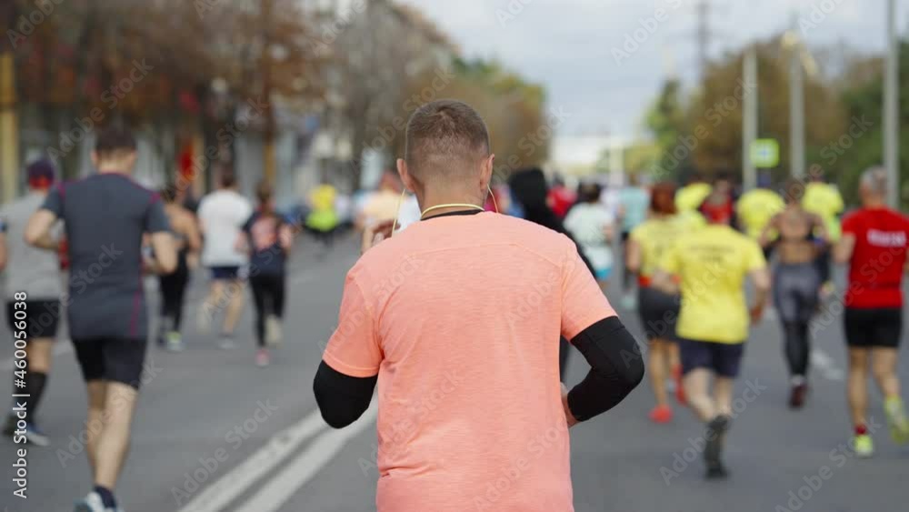 Slow motion people running marathon on empty city road, focus on man with earphones and bottle of water in hands. Following shot of sports event contestants. Concept of healthy lifestyle Slow motion people running marathon on empty city road, focus on man with earphones and bottle of water in hands. Following shot of sports event contestants. Concept of healthy lifestyle