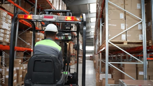 Back view of forklift driver riding between racks in storehouse with pallet of cardboard boxes to place on shelf. Male employee in helmet driving warehouse loader during work