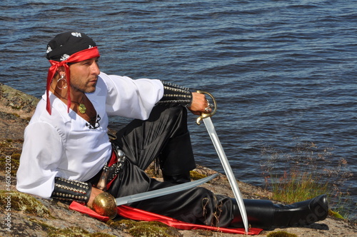 Side view man in pirate costume posing with swords and sitting near seashore 