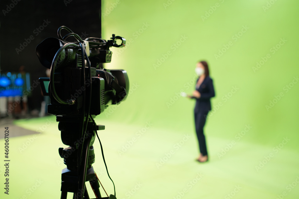 Selective focus of a video camera on a tripod are recording a blurred Asian female reporter in suit and wearing a face mask to protect Covid-19 standing talking on a green screen background in studio. Selective focus of a video camera on a tripod are recording a blurred Asian female reporter in suit and wearing a face mask to protect Covid-19 standing talking on a green screen background in studio.