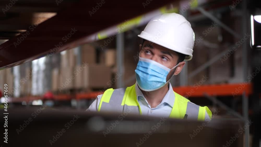 Bearded male supervisor in protective mask, hard hat and vest using tablet for stocktaking while standing at rack in storehouse. Focused inventory manager checking products stock in warehouse Bearded male supervisor in protective mask, hard hat and vest using tablet for stocktaking while standing at rack in storehouse. Focused inventory manager checking products stock in warehouse