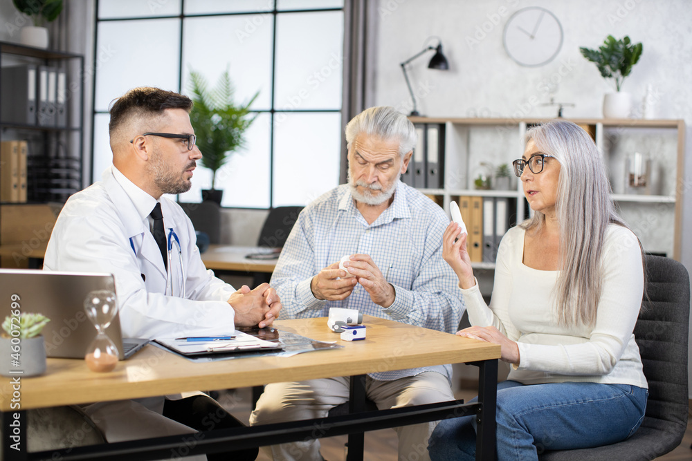 Caucasian medical worker white lab coat and eyewear prescribing treatment to mature couple that sitting at his cabinet. Aged man and woman visiting family doctor for health examination. Caucasian medical worker white lab coat and eyewear prescribing treatment to mature couple that sitting at his cabinet. Aged man and woman visiting family doctor for health examination.