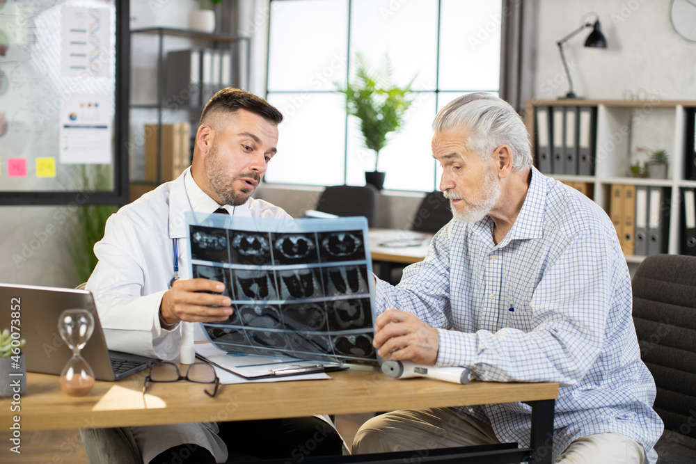 Caucasian medical worker in white lab coat showing x ray scan to senior male patient at private office. Aged man in casual wear listening attentively results of medical diagnostic.