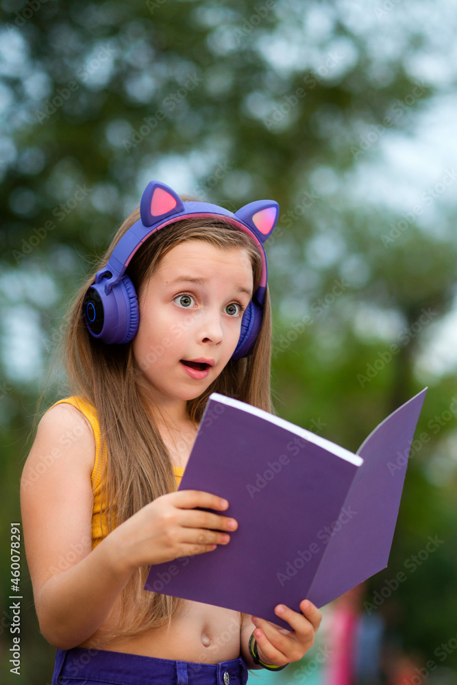 Surprised Primary School Girl Holding An Open Notebook. Preteen school girl with book on bench. Child with wireless headphones wears trendy clothes, chilling in park Surprised Primary School Girl Holding An Open Notebook. Preteen school girl with book on bench. Child with wireless headphones wears trendy clothes, chilling in park