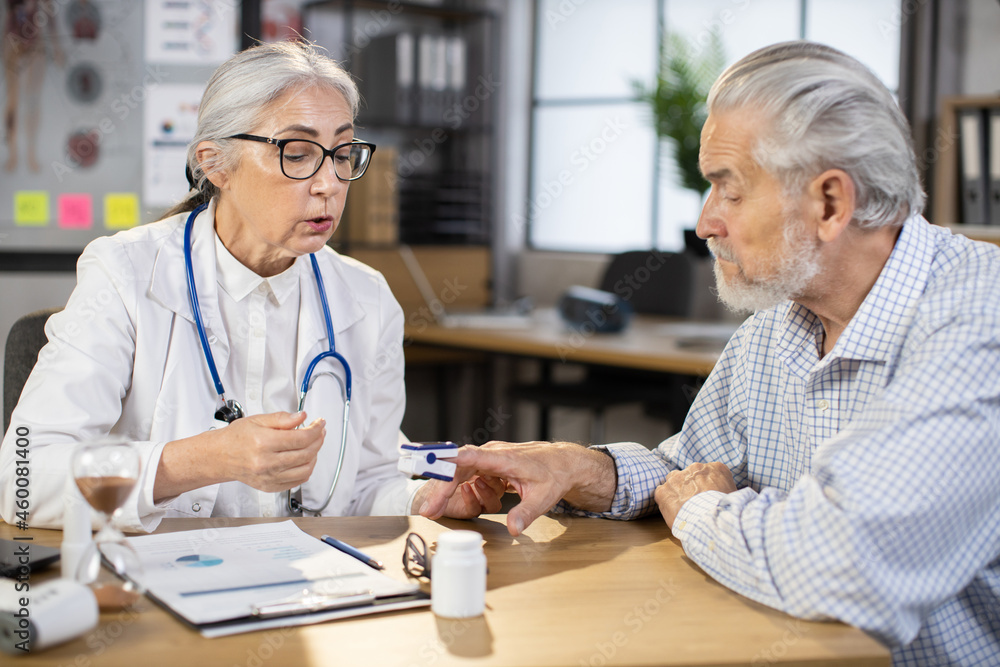 Mature female doctor in eyewear and uniform monitoring saturation of aged male patient using digital pulse oximeter. Concept of people, health care and treatment. Mature female doctor in eyewear and uniform monitoring saturation of aged male patient using digital pulse oximeter. Concept of people, health care and treatment.