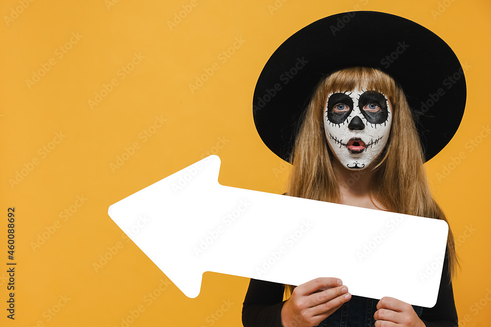 Scary bright alive girl child with Halloween makeup mask showing aside with white arrow wears big black hat, posing isolated over yellow color background studio with copy space for promotional content Scary bright alive girl child with Halloween makeup mask showing aside with white arrow wears big black hat, posing isolated over yellow color background studio with copy space for promotional content