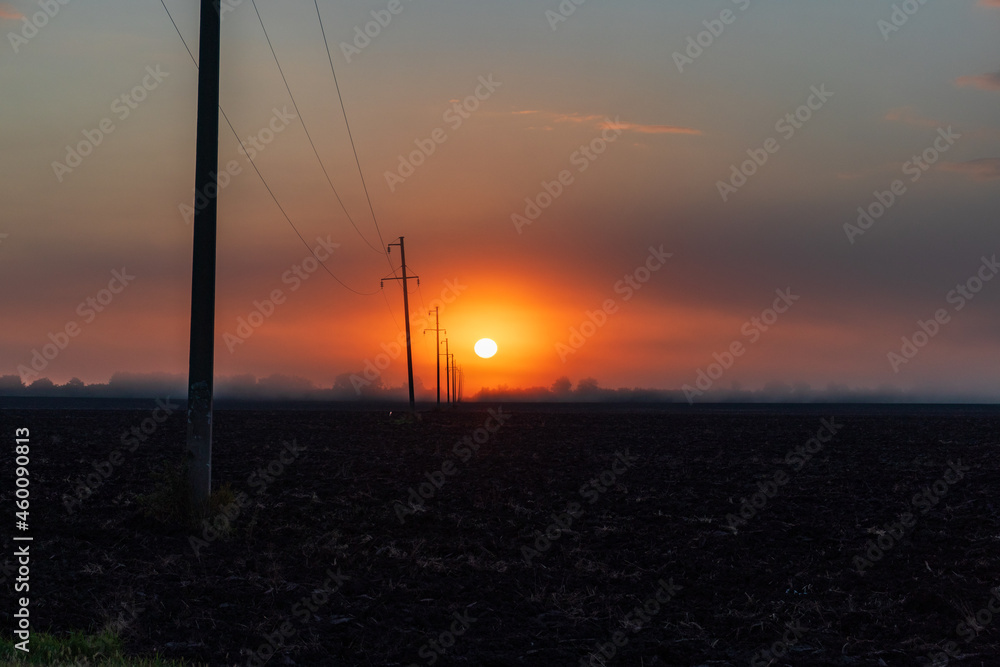 Silhouettes of utility poles carrying high voltage electricity alongside a road at a rural area. Sunset, sunrise image with cloudy orange red sky and depth of field with nobody in the frame.