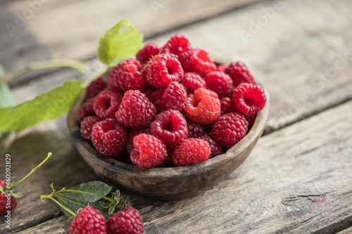 Appetizing juicy ripe raspberries in a bowl surrounded by green raspberry leaves on a wooden  background. Copy space