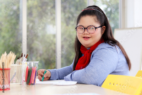 Portrait of happy girl having fun during study at school, child with down syndrome concentrate painting on paper in art classroom, kid with physical disability and intellectual education concept. photo