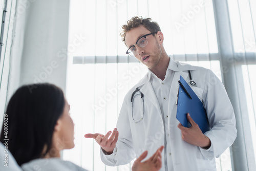 curly doctor in glasses holding clipboard and gesturing near patient