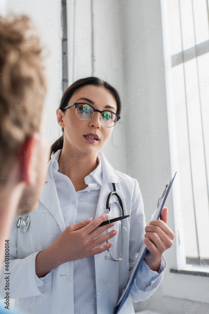 doctor in glasses holding pen and clipboard while looking at blurred patient doctor in glasses holding pen and clipboard while looking at blurred patient