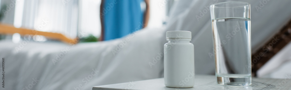 bottle with medication and glass of water on bedside table near blurred patient in hospital, banner