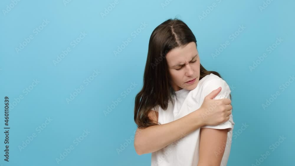 Unhappy sad tired young brunette woman has painful feelings in left shoulder, clenches teeth, wears white t-shirt, posing isolated over blue color background with copy space area for promotion content