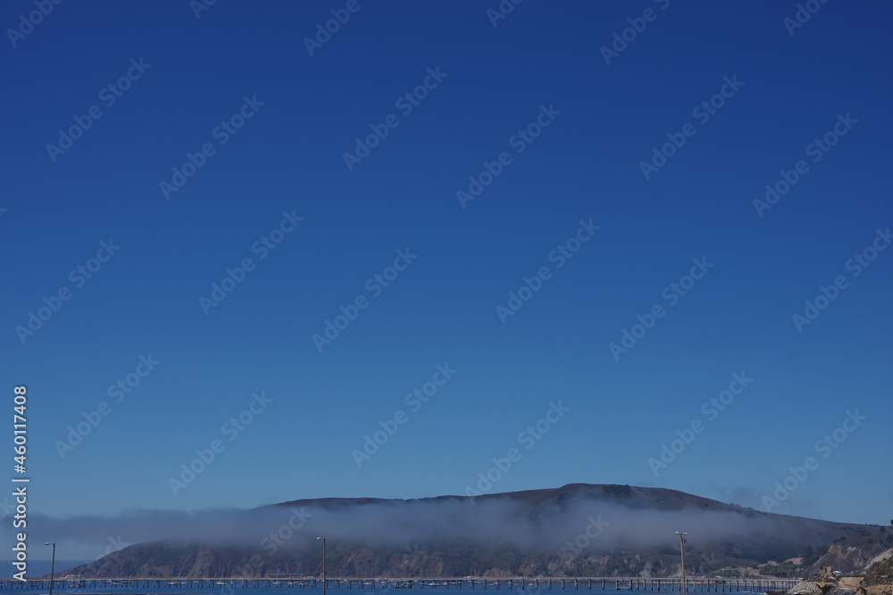 Panoramic view of the Port San Luis pier in California with a fog cloud, a mountain range behind and blue sky above seen from Avila beach
