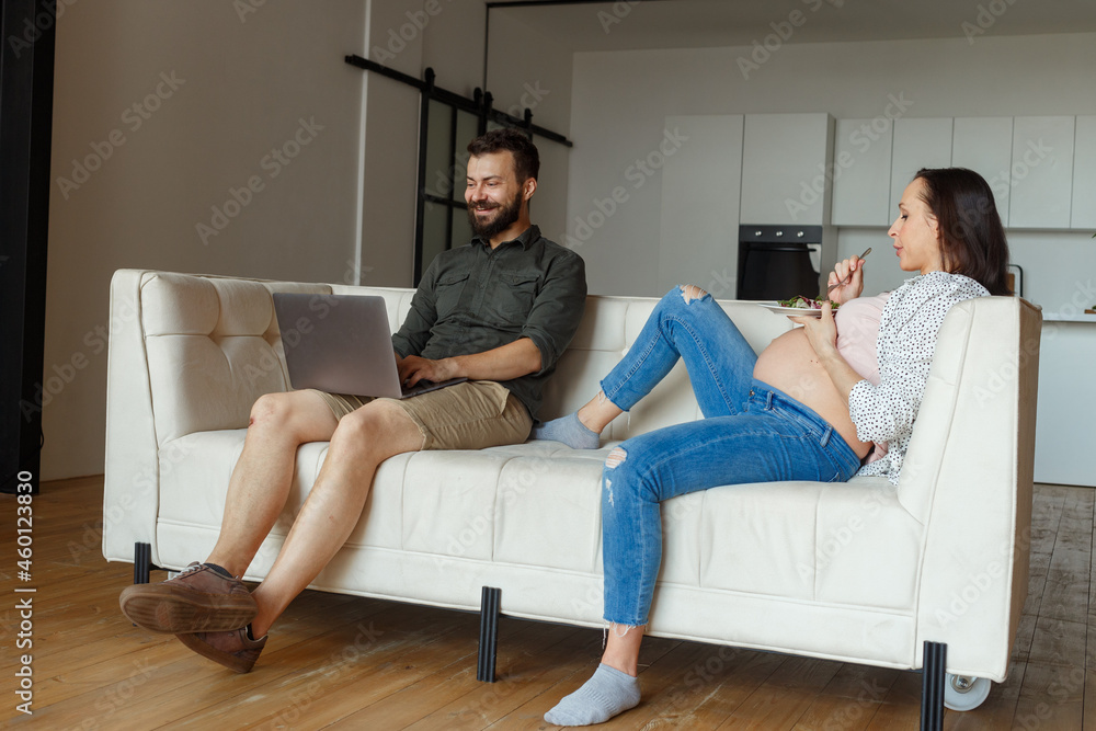 Young pregnant woman lies on white sofa holding plate with food near sitting husband with laptop against spacious light room Young pregnant woman lies on white sofa holding plate with food near sitting husband with laptop against spacious light room