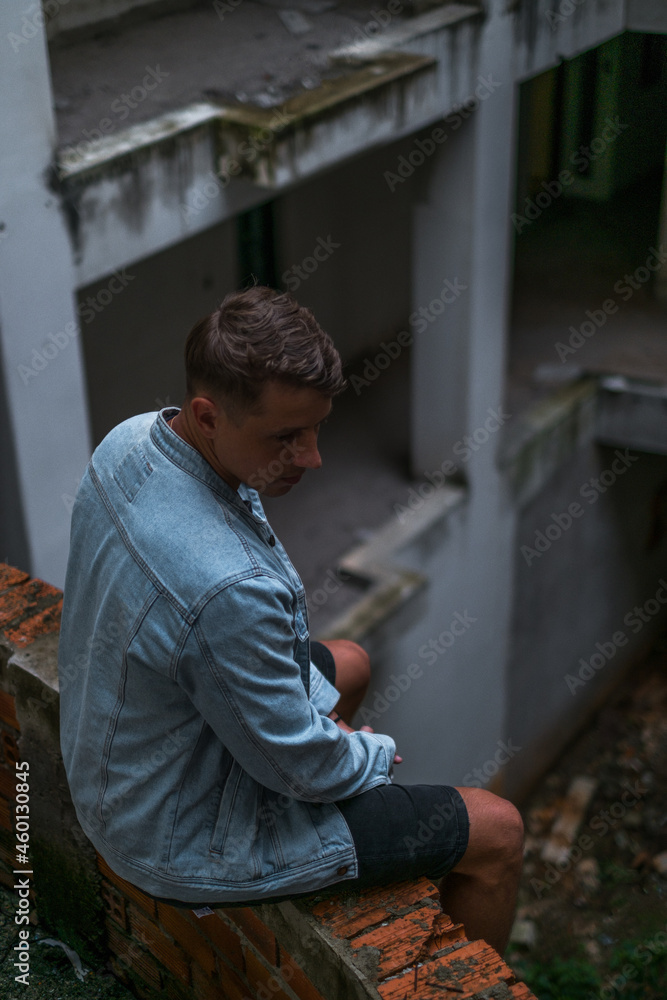 Portrait of young handsome Caucasian man in denim jacket, sitting on the abandoned construction site and looking down Portrait of young handsome Caucasian man in denim jacket, sitting on the abandoned construction site and looking down