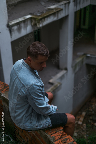 Portrait of young handsome Caucasian man in denim jacket, sitting on the abandoned construction site and looking down 