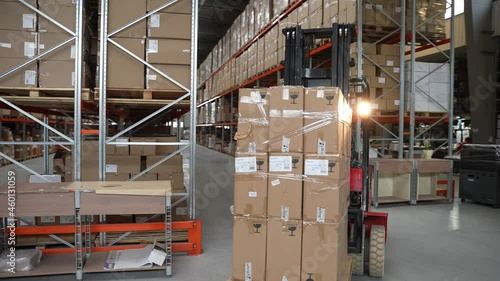 Professional forklift driver moving huge pallet of cardboard boxes while riding between rows of racks in storehouse. Male worker in hard hat driving loader with goods around warehouse