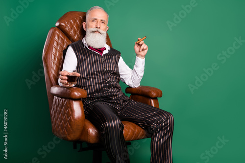 Photo of old noble aristocrat sit armchair drink whiskey enjoy cigar wear striped suit on green color background
