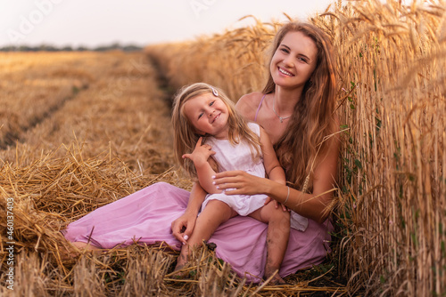 Mother and little daughter have fun sitting by the golden wheat field. Young woman tickles the girl