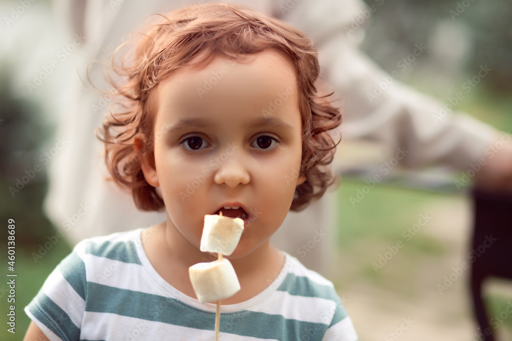 Little curly girl eating roasted marshmalloy outdoor in a camp. Travel, hike, vacation, camping concept.