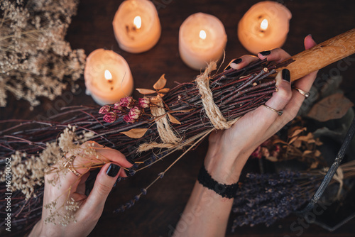 Wiccan witch decorating a DIY besom broom for Samhain celebration. Hand made broom on a dark wooden table with white lit candles and colorful dried flowers and herbs in the background