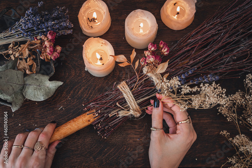 Wiccan witch decorating a DIY besom broom for Samhain celebration. Hand made broom on a dark wooden table with white lit candles and colorful dried flowers and herbs in the background photo