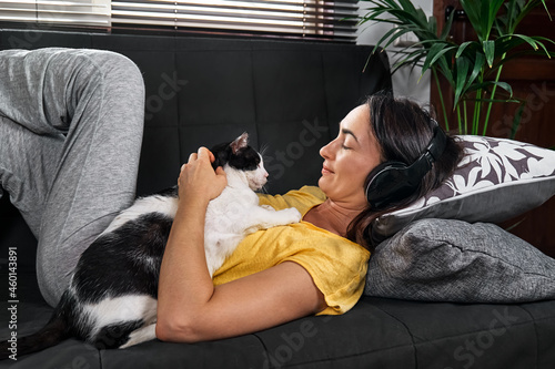 Woman cuddles her funny sleeping cat next to her on the sofa while listening music with headphones at home. Woman listening music with smartphone. Gadgets in the life. ​People on the couch photo