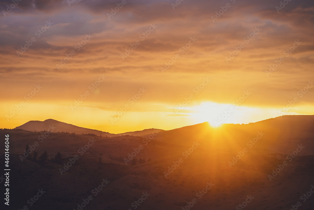 Atmospheric landscape with silhouettes of mountains with trees on background of dawn sky with sun circle and orange sun rays. Colorful nature scenery with sunset or sunrise of illuminating color.