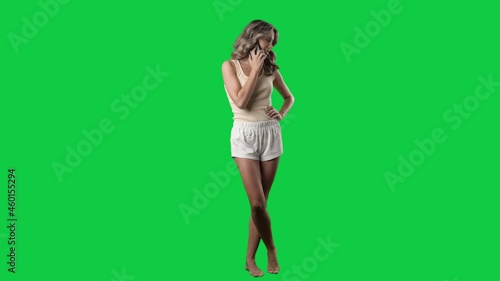 Relaxed candid happy young woman in pajamas talking and laughing on cellphone. Full body on green screen chroma key background