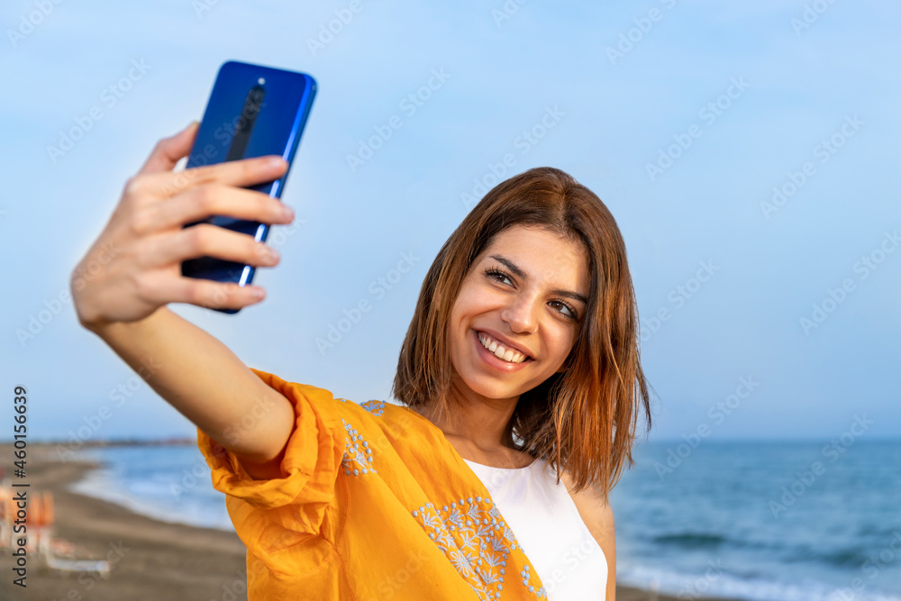 Smiling young brunette taking a selfie on the beach. The sunset light enhances the happiness on her face. Quiet beach and sea in the background.