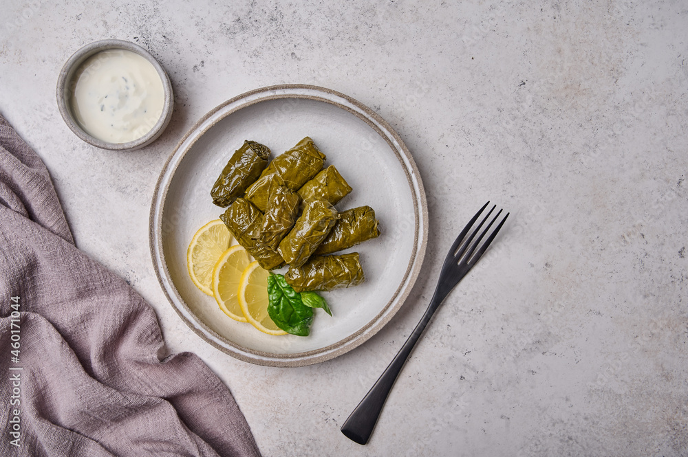Flat lay dolma, stuffed grape leaves with rice, meat, lemon in light plate with with sour cream, black fork and linen napkin
