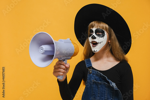 Portrait of spooky child girl with Halloween makeup mask screaming in megaphone looking aside, wears big black hat, posing isolated over yellow color background wall in studio. Party holiday concept