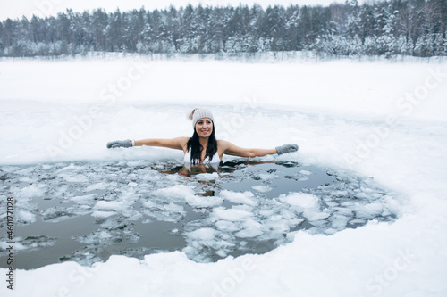 Winter cold swimming. Woman in frozen lake ice hole. Swimmers wellness in icy water. How to swim in cold water. Beautiful young female in zen meditation. Hat and gloves swimming clothes. Nature lake