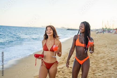 Cheerful girlfriends with drinks and sound speaker on beach