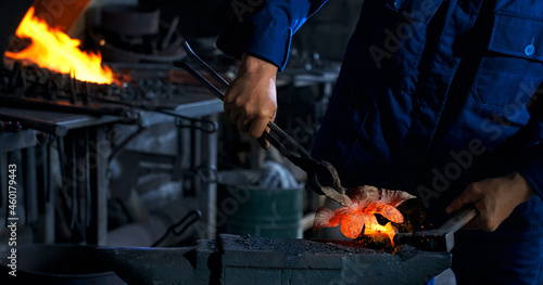 Man in blue uniform using hammer and pliers while working with molten metal at forge. Professional blacksmith processing hot steel at workshop.