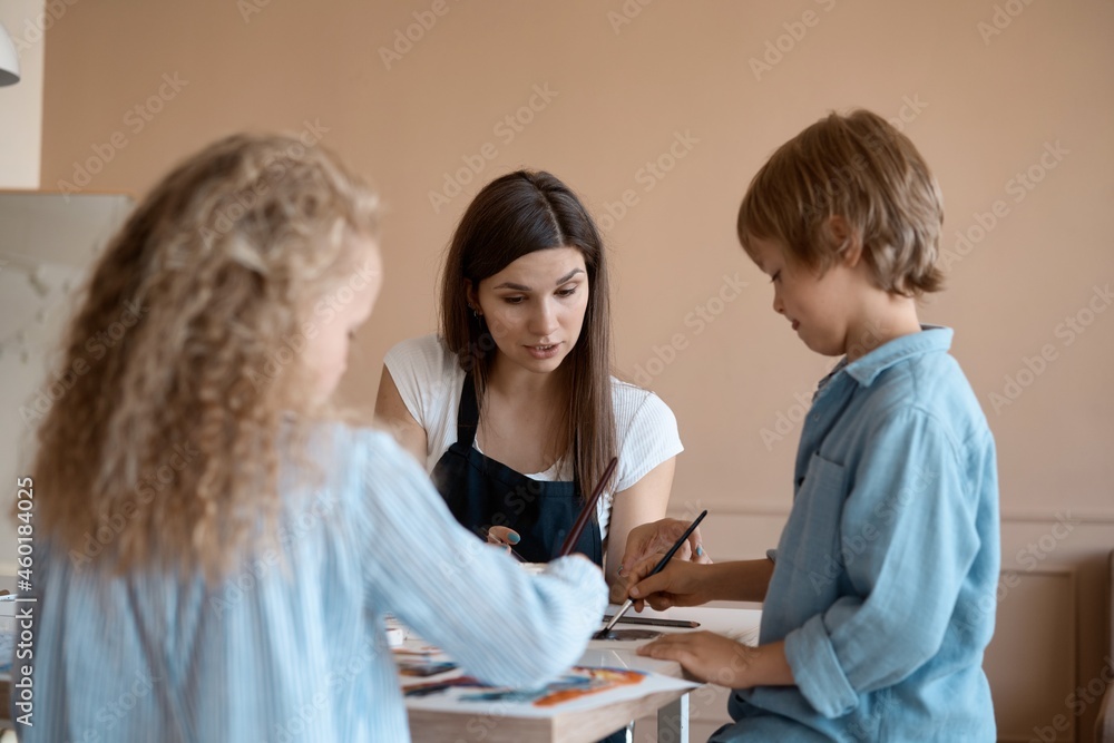 Group of kids and their teacher leaning over table with watercolor paints and painting self-made clay items. Children with teacher in art class. Group of kids and their teacher leaning over table with watercolor paints and painting self-made clay items. Children with teacher in art class.