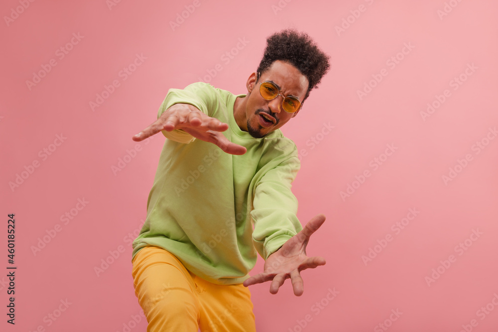 Attractive young American man reaches with both hands for camera against pink background. Basketball player made decisive shot in championship in gorgeous outfit of green long sleeves and yellow jeans