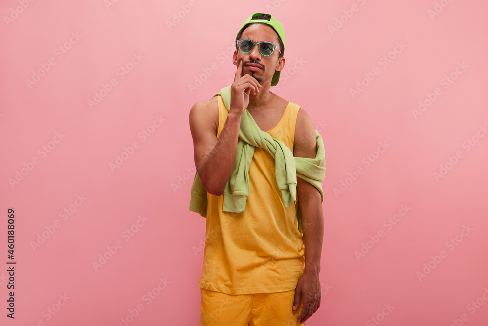 Student against pink wall express pensive emotion, touching chin, looks aside. Medium shot of thoughtful impressive young brown-skinned man with green eyewear, dressed up colourful. Student against pink wall express pensive emotion, touching chin, looks aside. Medium shot of thoughtful impressive young brown-skinned man with green eyewear, dressed up colourful.