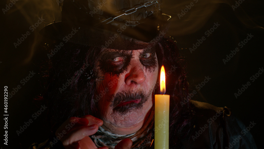 Fototapeta premium Frightening senior woman with Halloween witch makeup looking at candle, conjure, hex, wiz. Horror theme. Sinister sorcerer lady on studio black background. Voodoo magical creepy rituals with candle