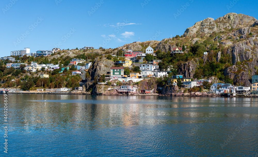 The hillside of St. John's Harbour, on a sunny day, under blue sky and white clouds. The colorful wooden houses are scattered along the hillside with the blue ocean in the foreground. 