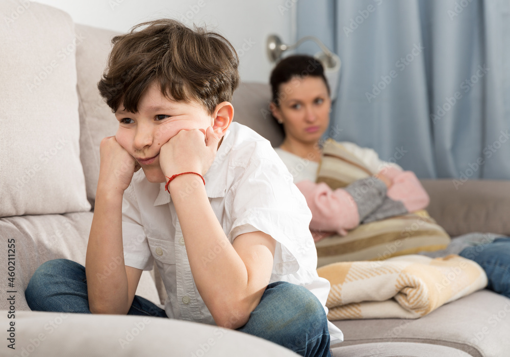 Upset preteen boy sitting on sofa and looking away after quarrel with mother..