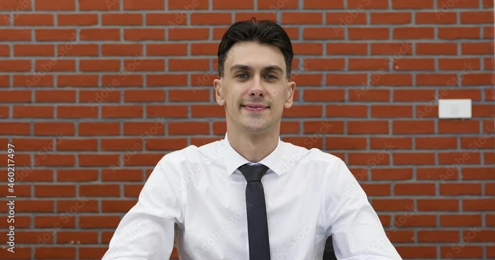 Young adult businessman in casual white shirt sitting in front of red brick wall, raise hand to camera for greeting to speaker in online conference and stay listening. Young adult businessman in casual white shirt sitting in front of red brick wall, raise hand to camera for greeting to speaker in online conference and stay listening.