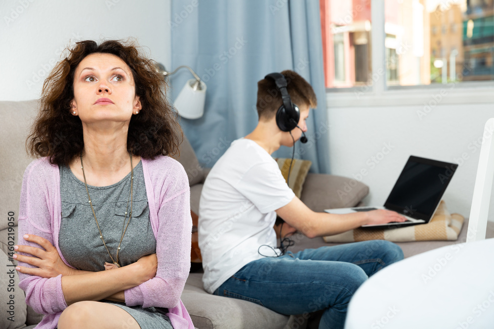 Obraz premium Portrait of upset woman sitting at home on background with her son playing on laptop. Computer addiction concept