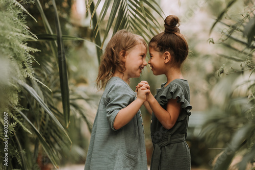 Two cute smiling little girls belonging to different races, in linen clothes, holding hands and walking in the botanical garden. Children explore tropical plants and flowers in the greenhouse.